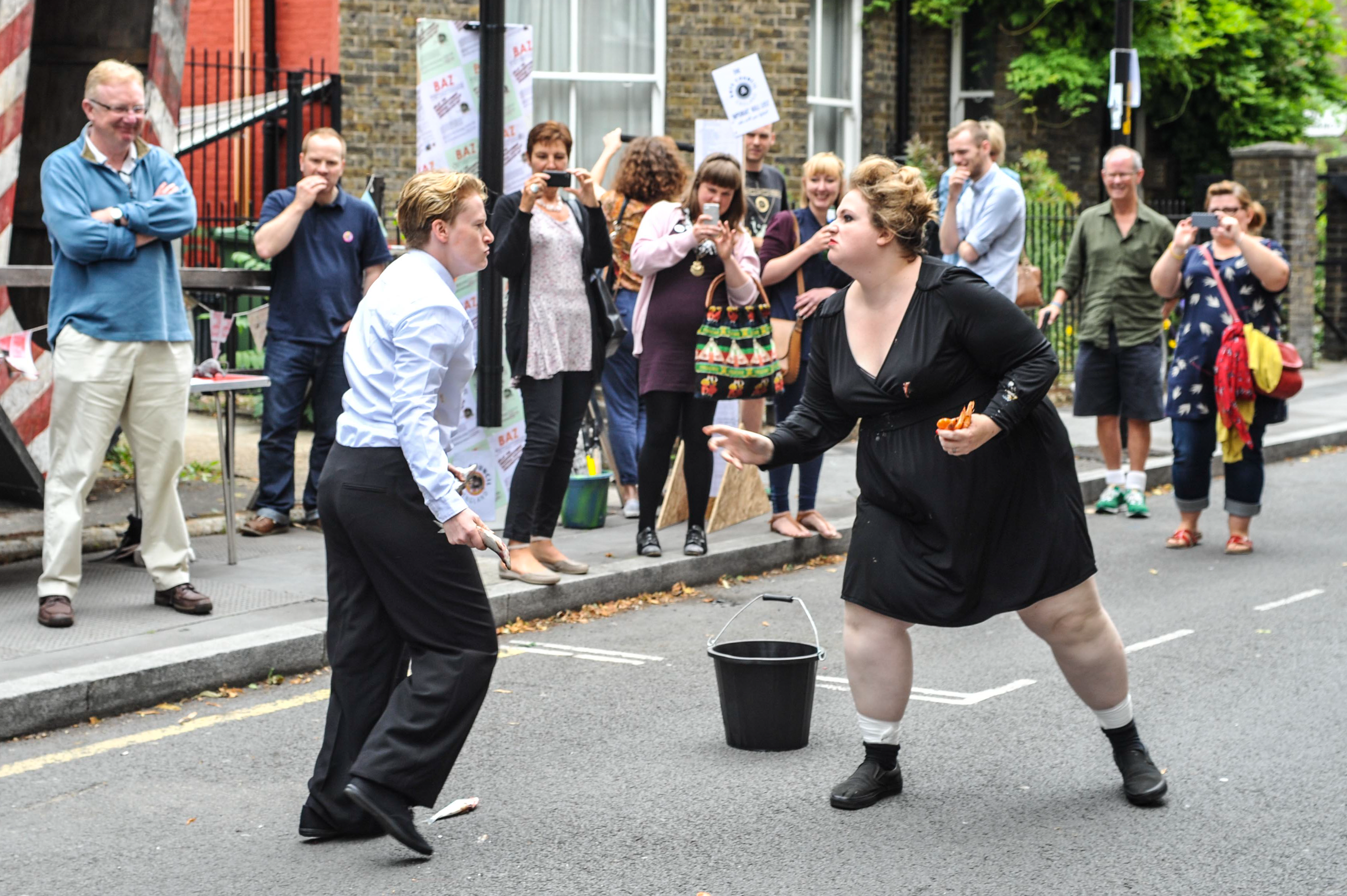 Two female bodies are stood in the middle of the street, they are both side on in confrontation. The individual on the left is wearing a black dress and is holding shellfish.  The indicudal on the left is wearing a white shirt, black trousers and is holding a small fish. On the floor between them is a black bucket. Behind them in the background stand onlookers.